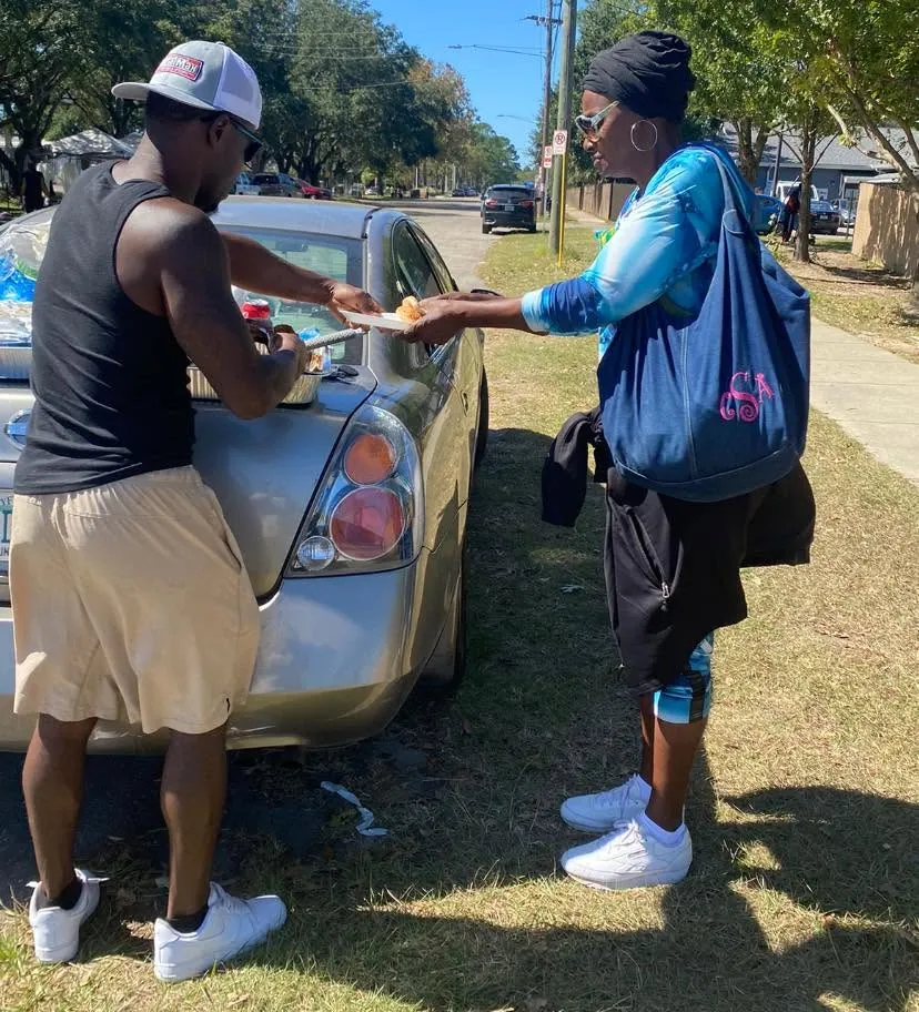 A House of Care volunteer handing a plate of food to a neighbor beside a parked car